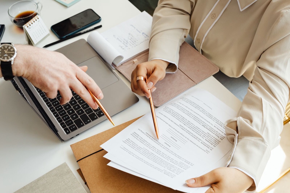 Two people's hands with pens over printed pages beside a laptop