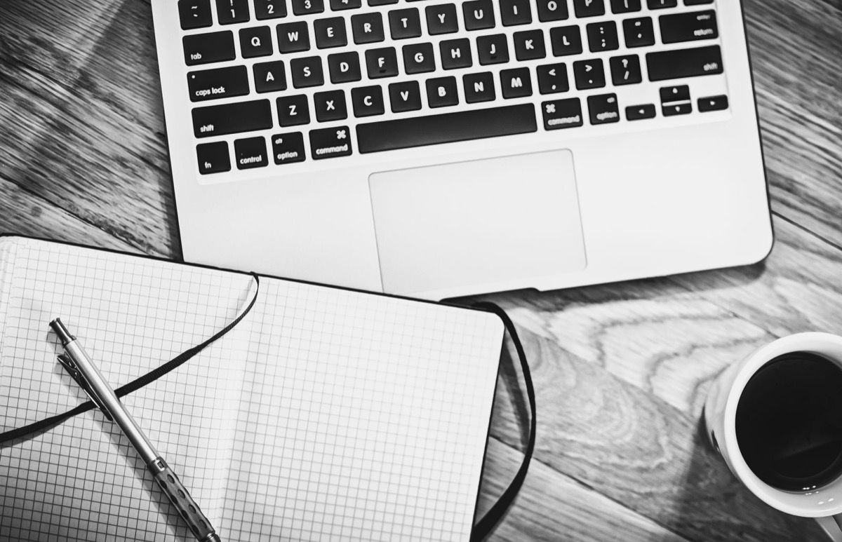 A laptop beside an open notebook, a pen and a cup of coffee