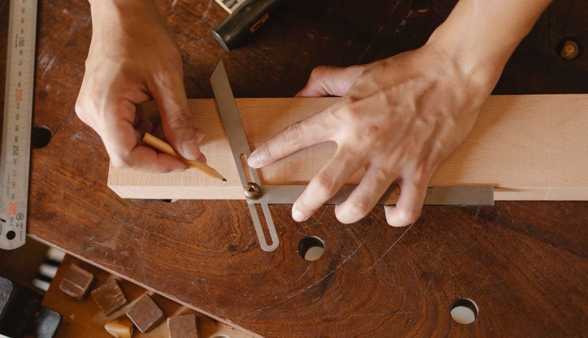 A craftsperson's hands marking a piece of wood with a bevel gauge and a pencil